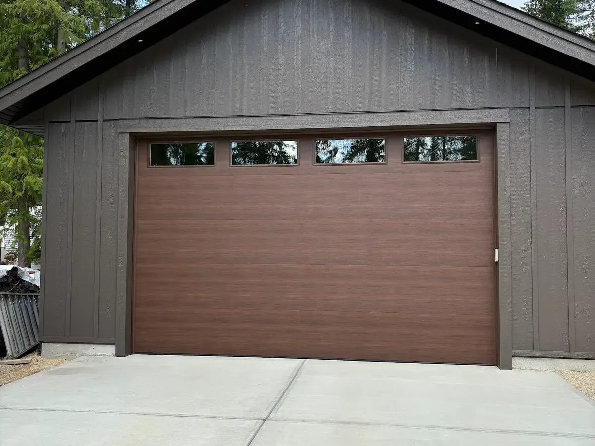 A walnut woodgrain garage door with four top windows on a dark board-and-batten detached garage