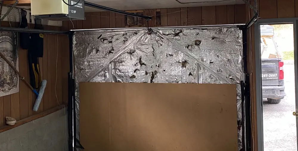 Inside a single garage, the door opening covered with torn foil insulation and a sheet of hardboard