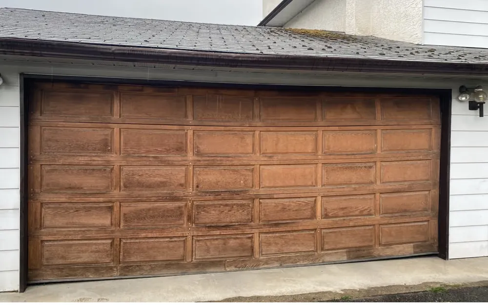 A weathered brown raised-panel double garage door on a white-sided home