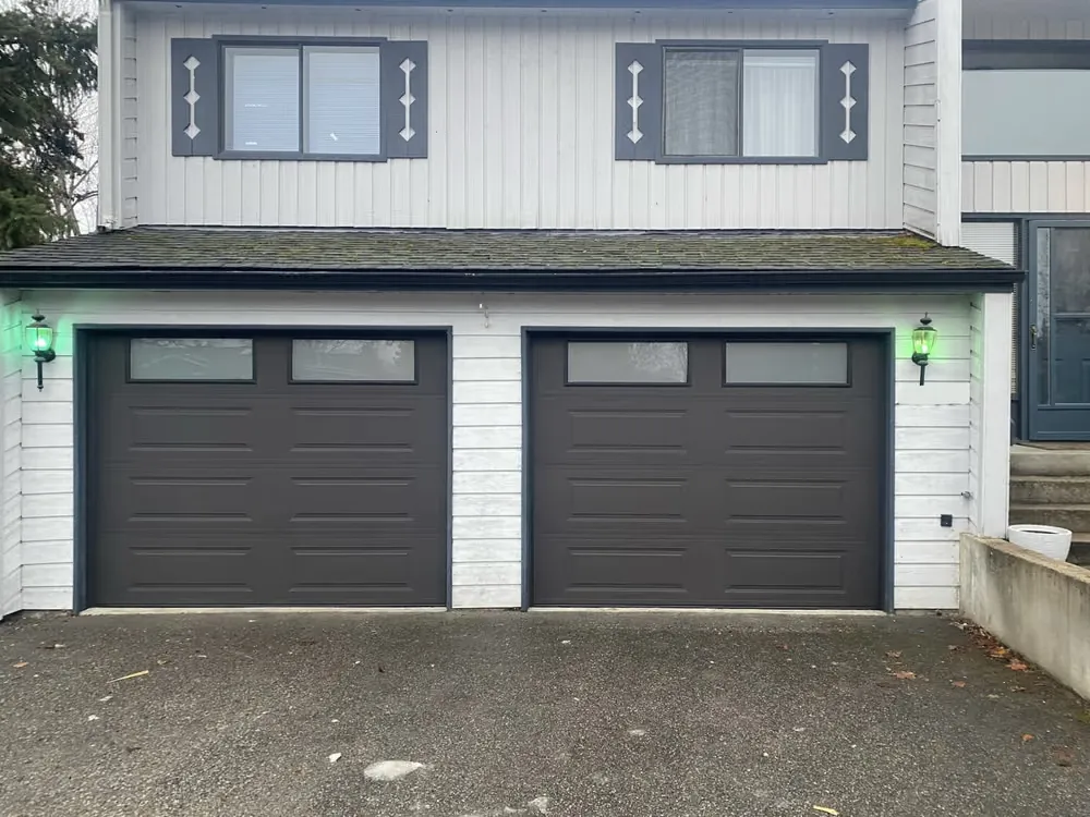 Two charcoal garage doors with frosted top windows between wall lanterns on the same home