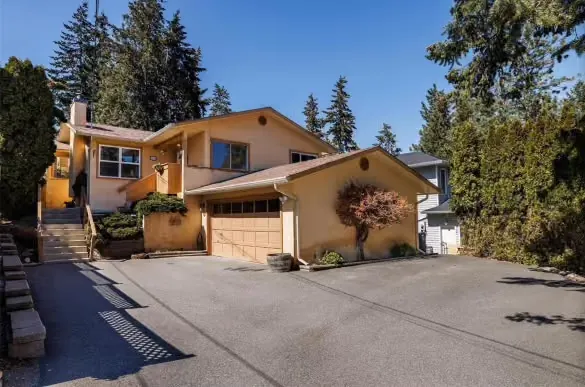 A tan stucco home with a beige double garage door at the end of a wide driveway, tall evergreens behind