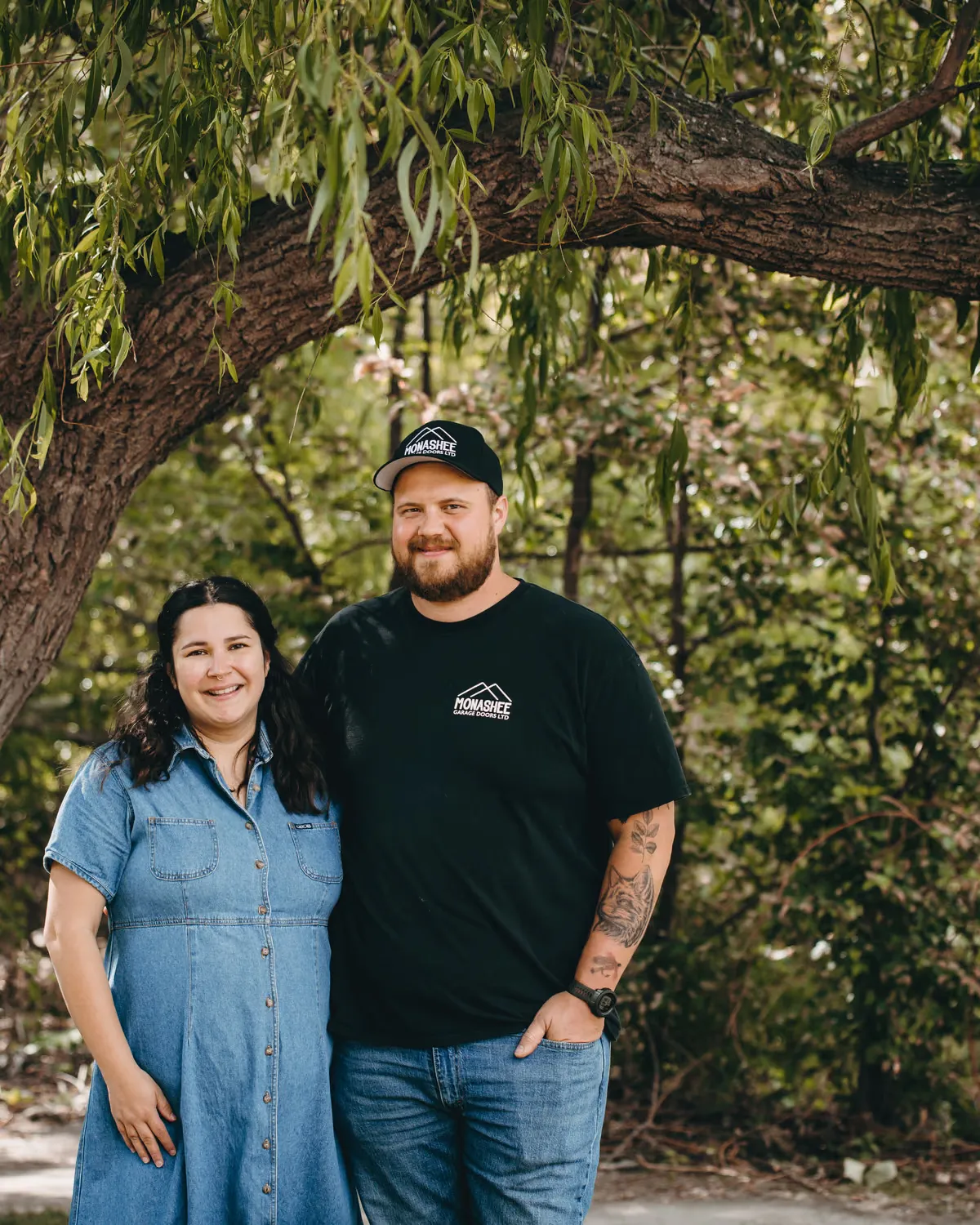 Josh and Haley Strother in Monashee Garage Doors gear, standing under a willow tree