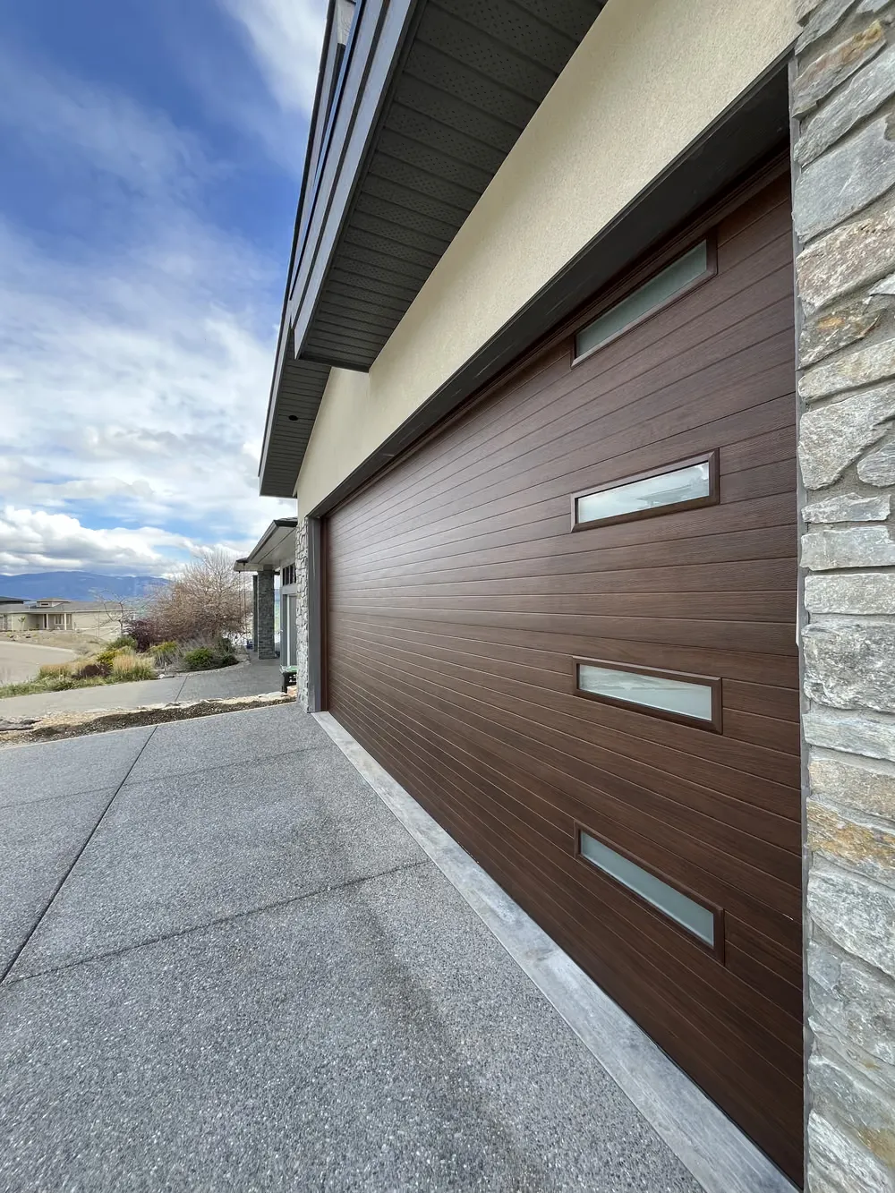 A walnut wood-look garage door beside a stone-clad wall, photographed down the driveway