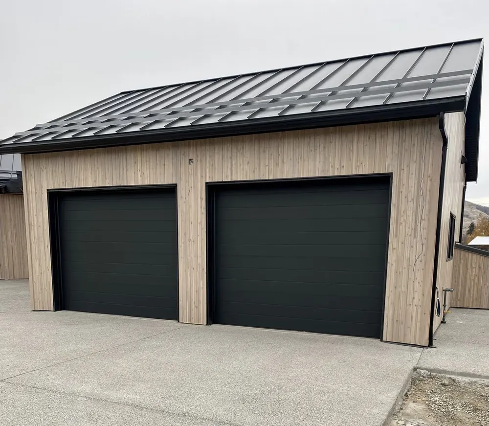 Two black garage doors on a shop clad in pale wood under a standing-seam metal roof