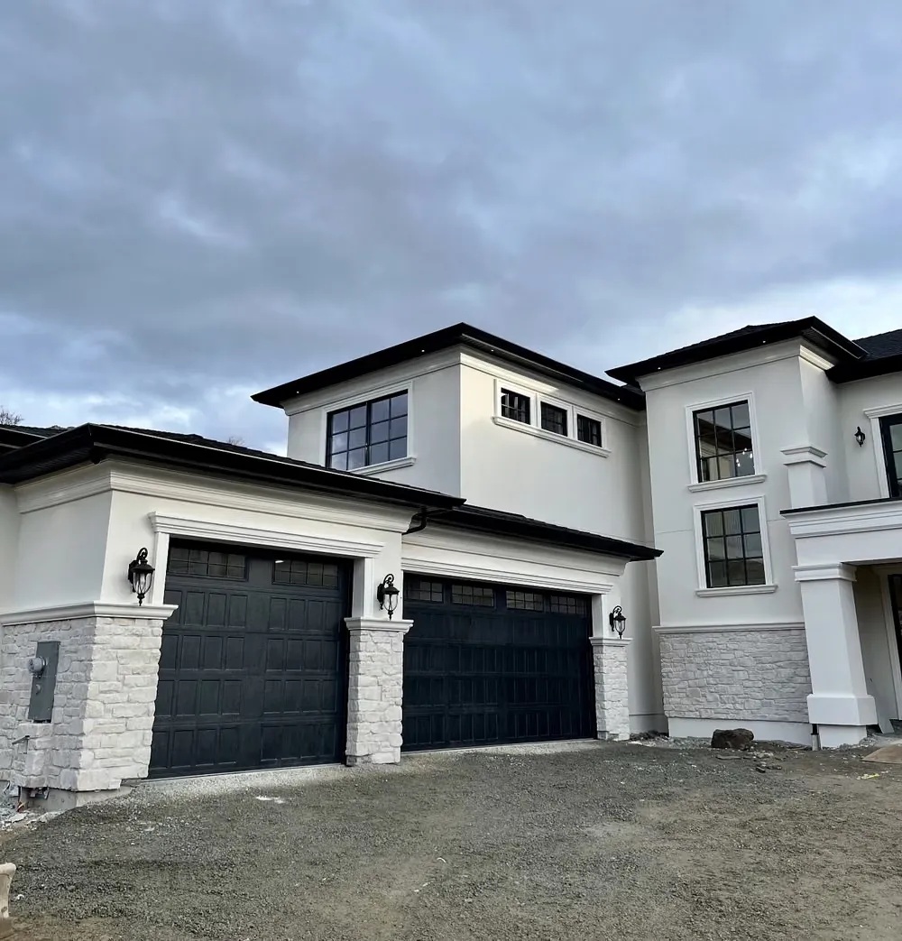 Two black garage doors between stone piers on a white stucco home nearing completion