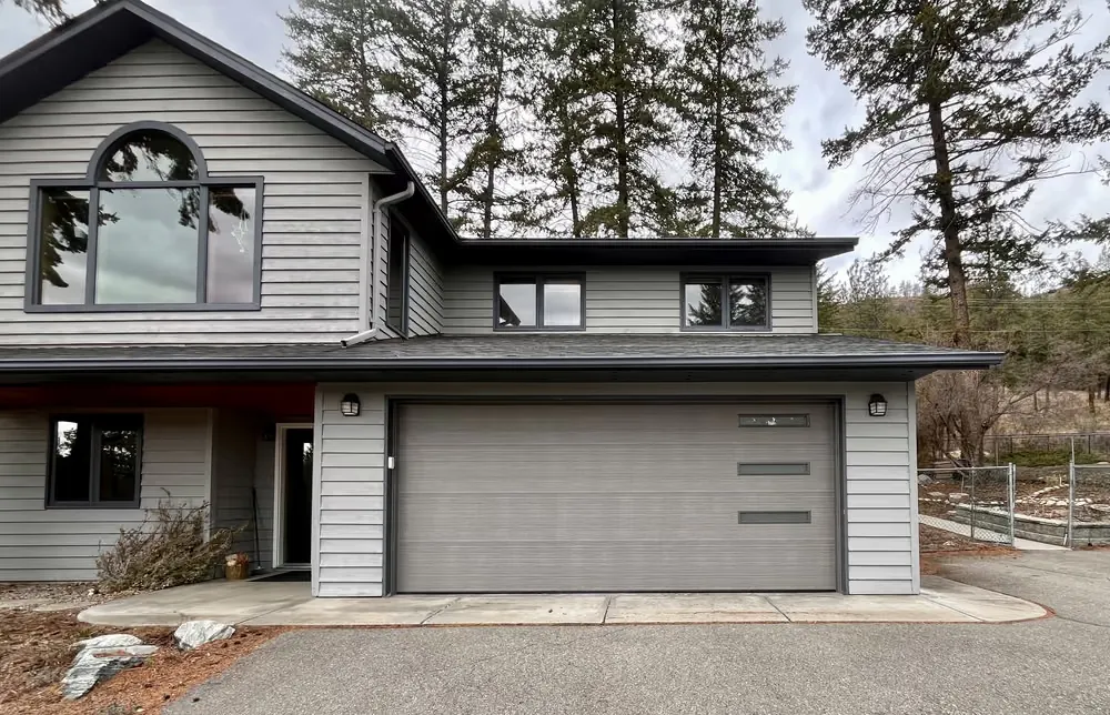 A taupe garage door with a frosted window strip on a grey two-storey home with an arched feature window