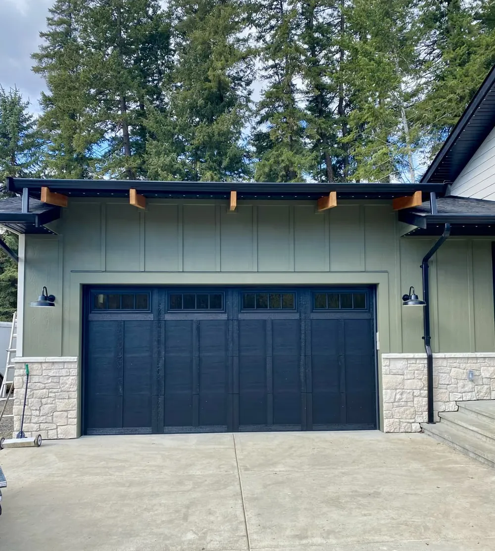 A black carriage-style garage door with square windows on a sage-green home with a stone skirt