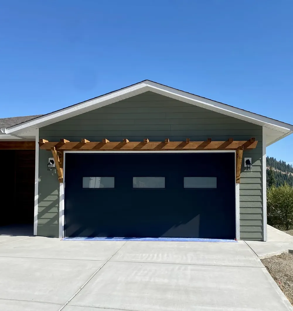 A dark garage door with a row of windows under a cedar pergola on a sage-green home