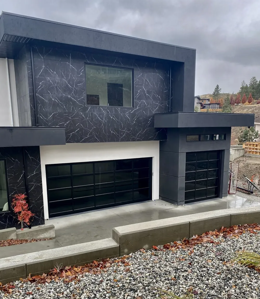 Two black full-view glass garage doors on a modern home clad in dark marble-look panels