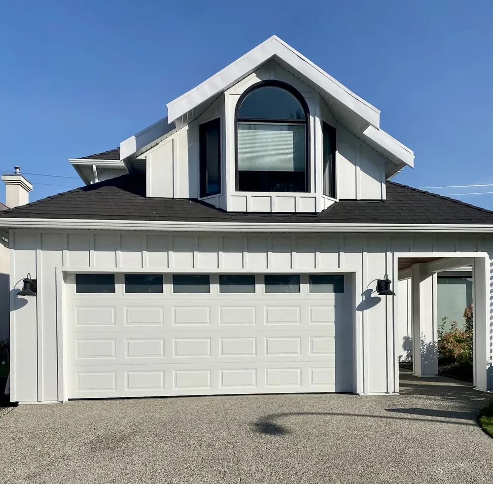 A white raised-panel double garage door on a white home with a black roof and an arched dormer