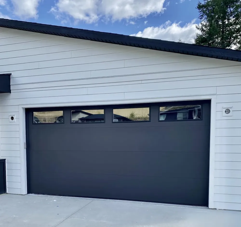 A black garage door with a row of top windows on a white shop with horizontal siding