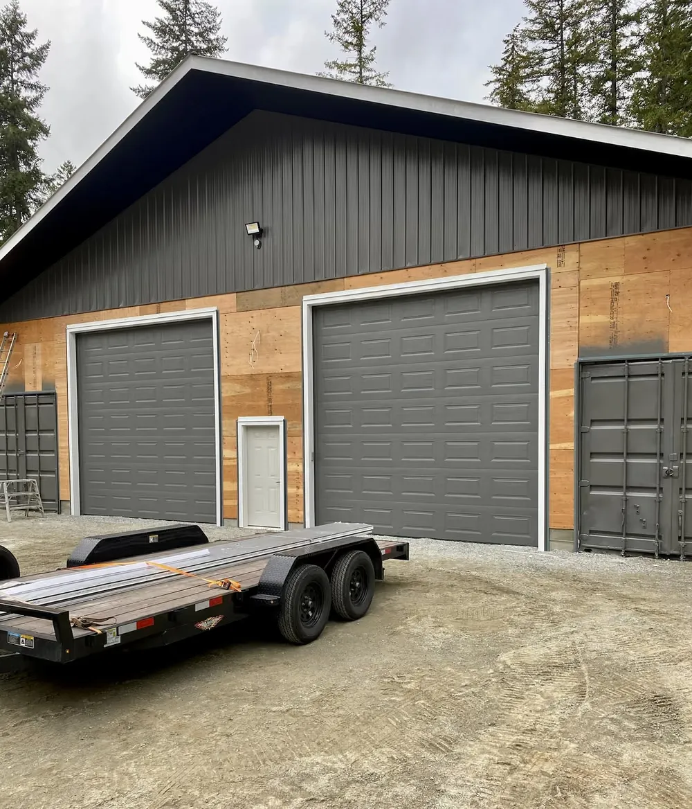Two dark grey garage doors on a shop with cedar and steel siding, a flat-deck trailer parked outside