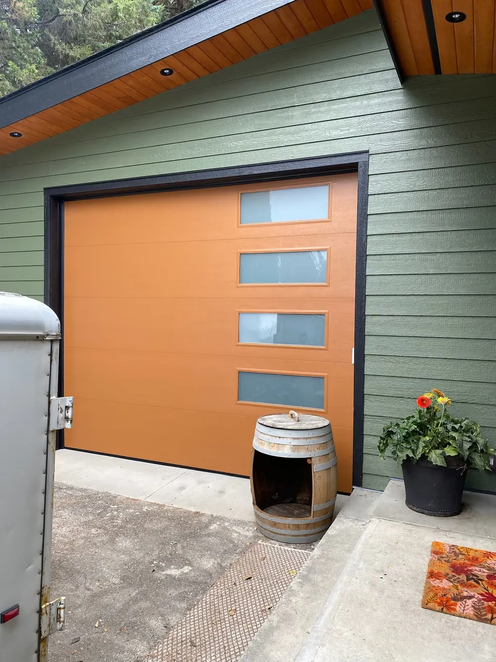 An orange garage door with a column of frosted windows on a green lap-sided building, a wine barrel beside it