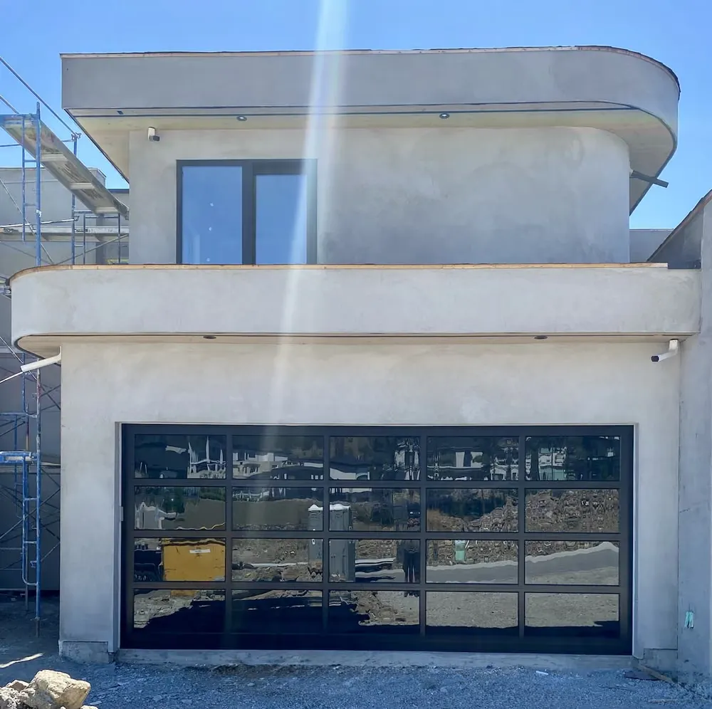 A full-view glass garage door on a curved white stucco build still ringed with scaffolding