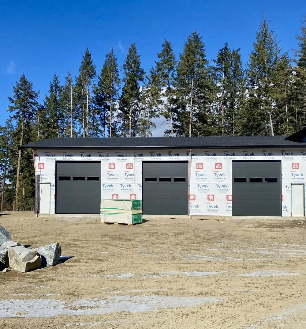 Three black garage doors on a shop still wrapped in house wrap, with fresh gravel out front