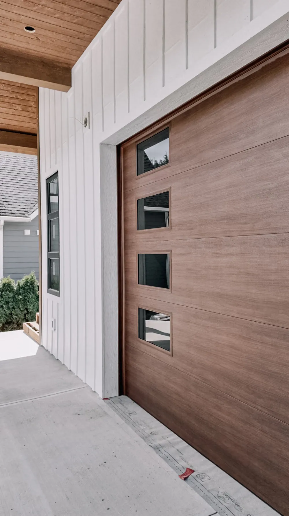 A wood-look garage door with a column of narrow windows on a white board-and-batten home with cedar soffits