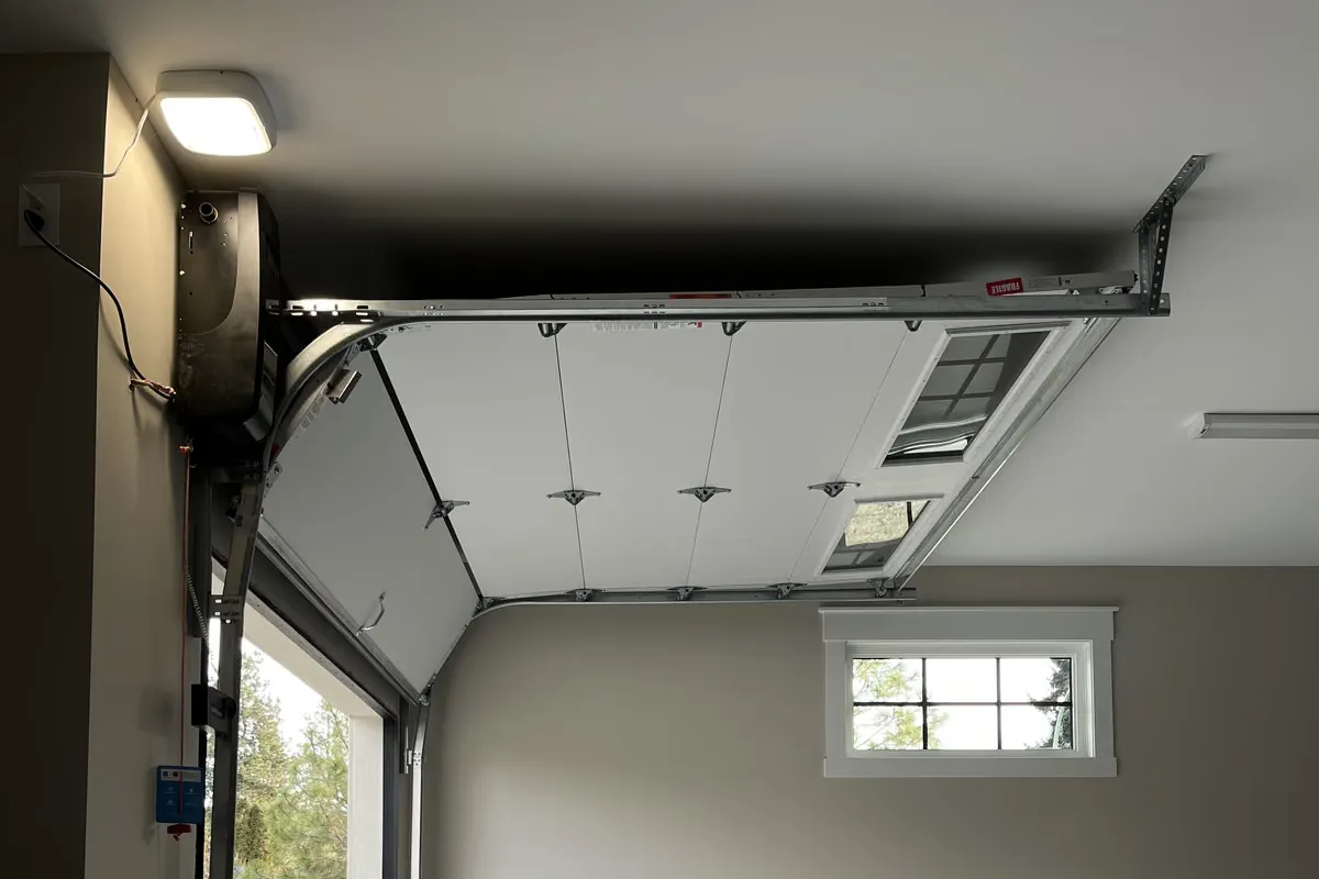 A white garage door rolled up along its ceiling tracks beside a wall-mounted opener and light